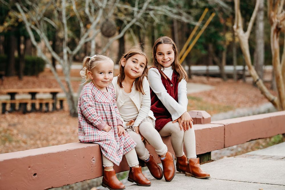 Three sisters in coordinating fall outfits sit together smiling on a wooden bench in a park.
