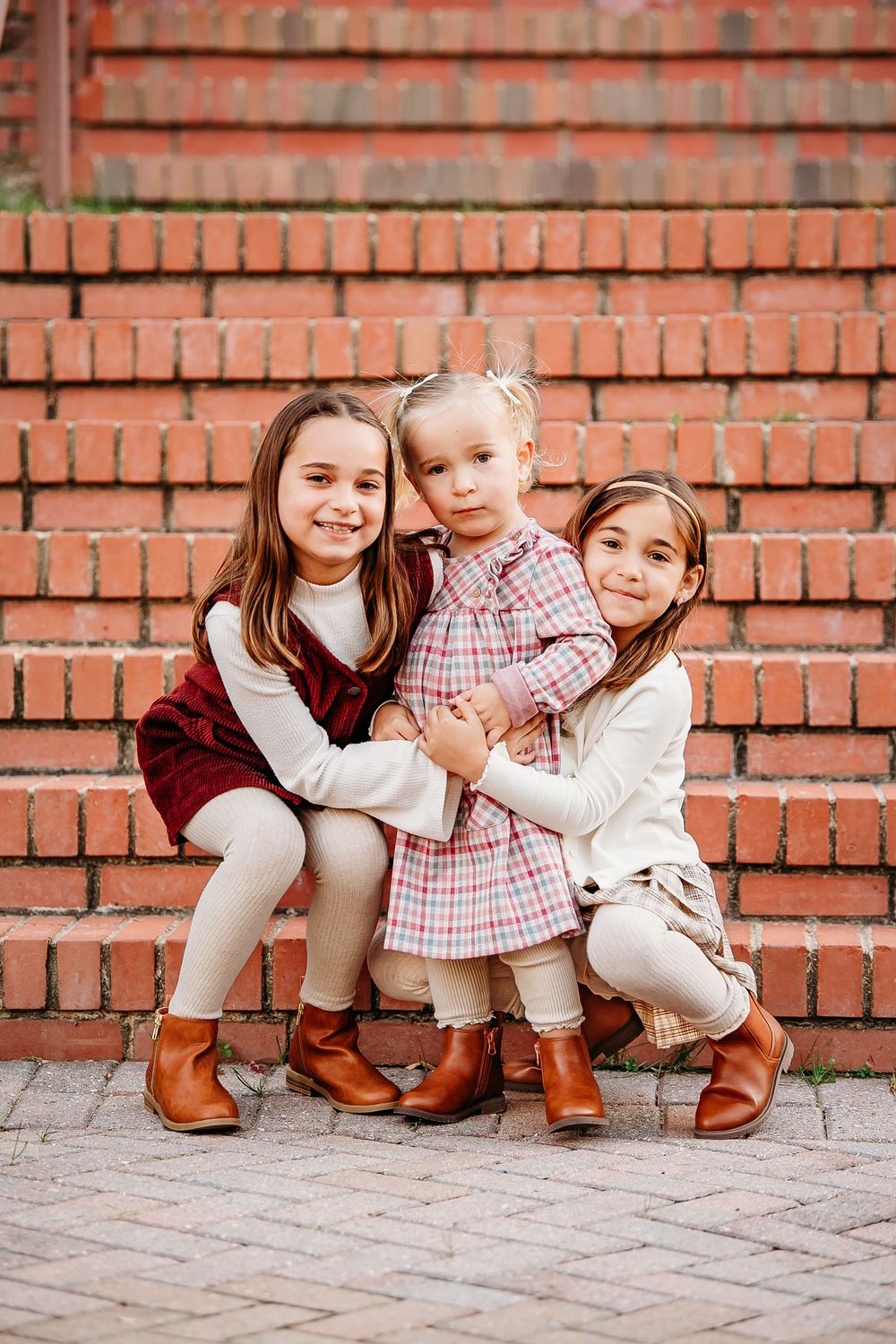 Three young sisters in coordinating fall outfits hug together in front of a red brick wall.