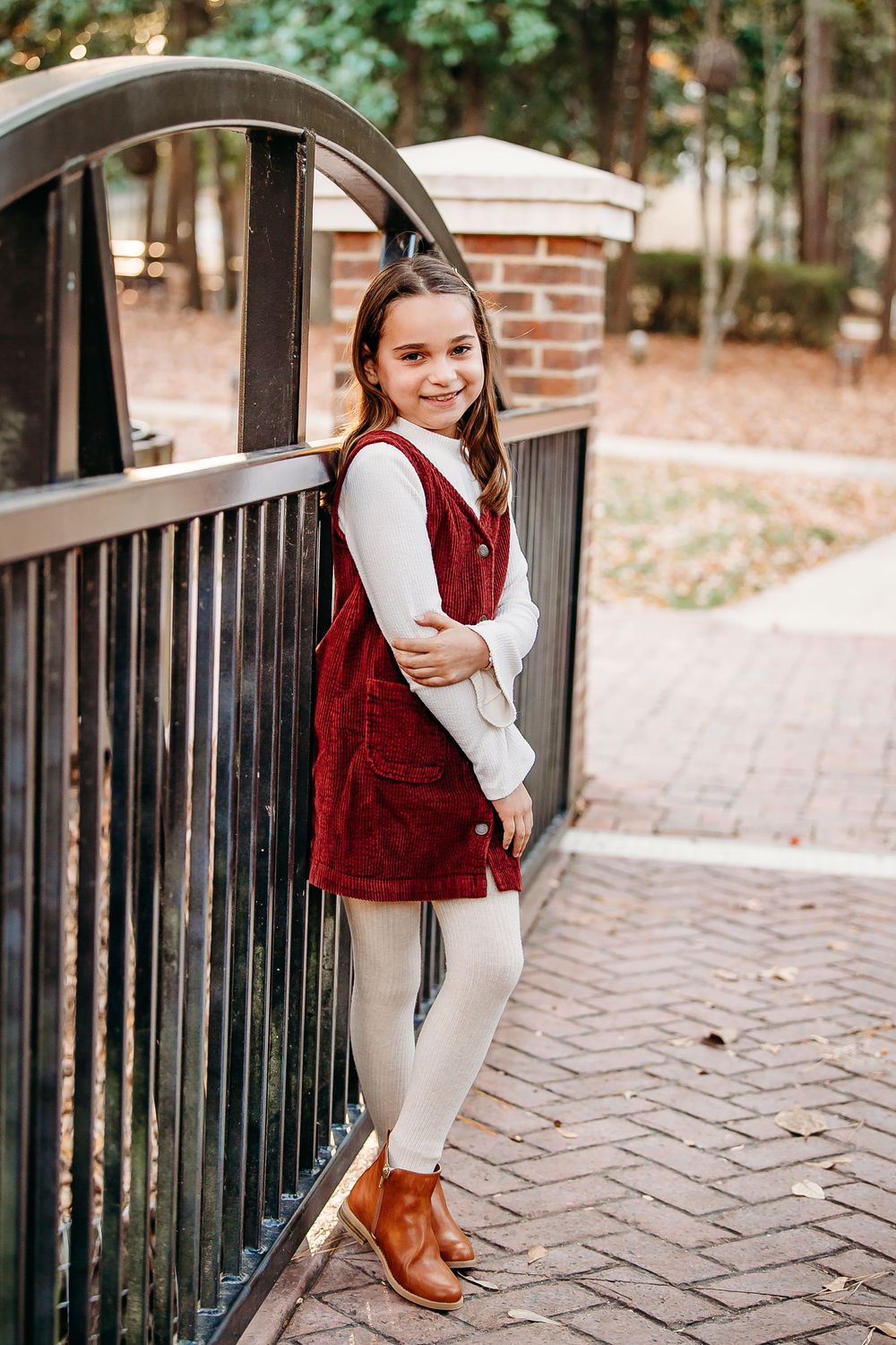 A smiling young girl in a red corduroy dress and brown boots leans against a metal fence in a park.