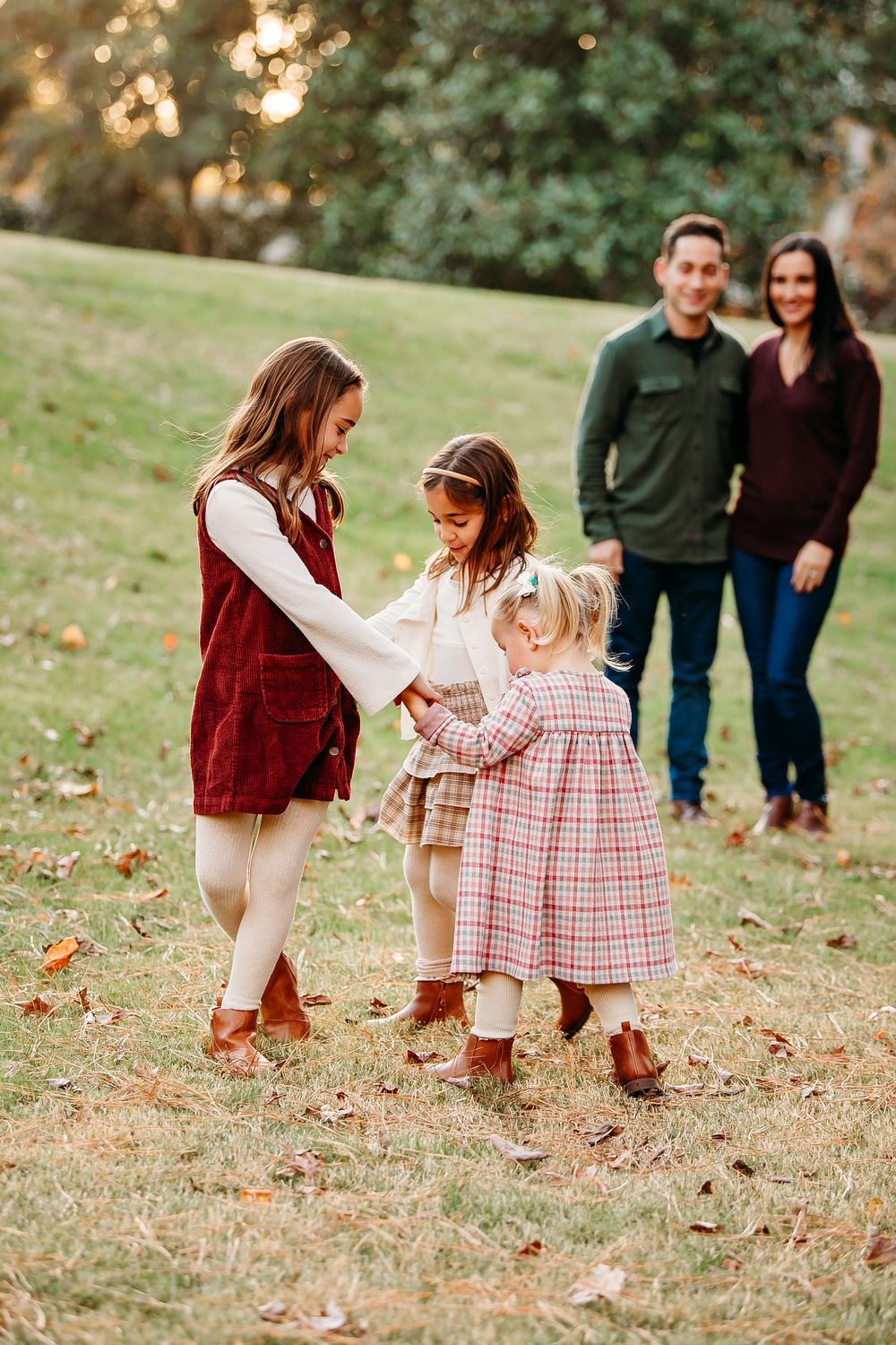 Two parents watch their three young daughters hold hands and play together on a grassy lawn.