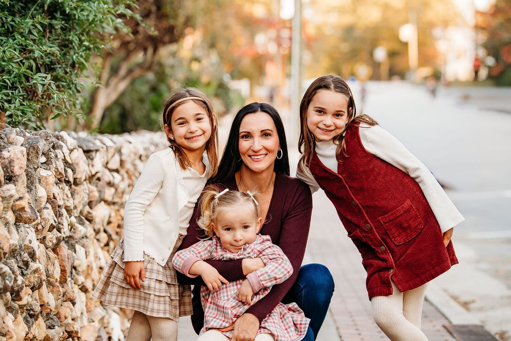 A mother crouches with her three daughters for a portrait along a sunny outdoor walkway.