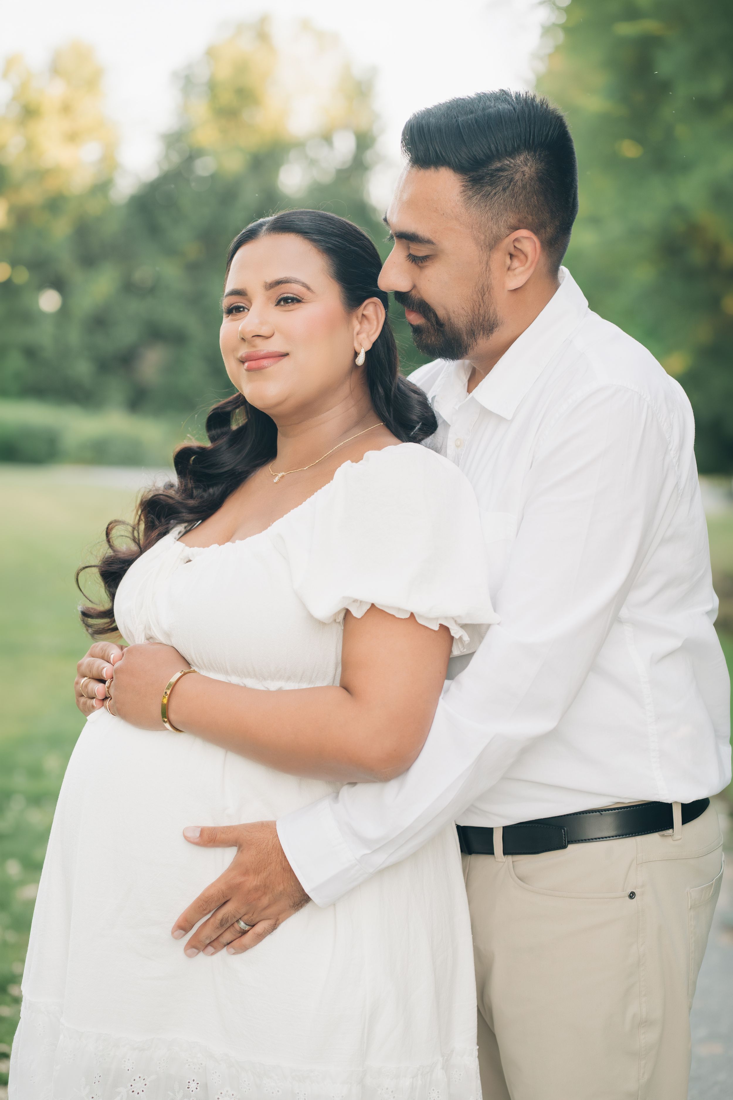 Pregnant woman in white dress embraced by partner outdoors in lush green park during golden hour maternity photo session.