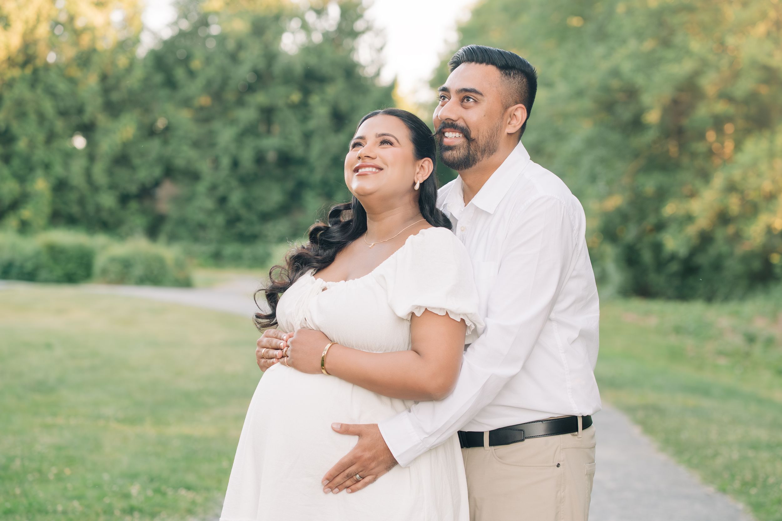 Pregnant couple embracing outdoors in a park, woman in white maternity dress, man in white shirt, lush green trees background.