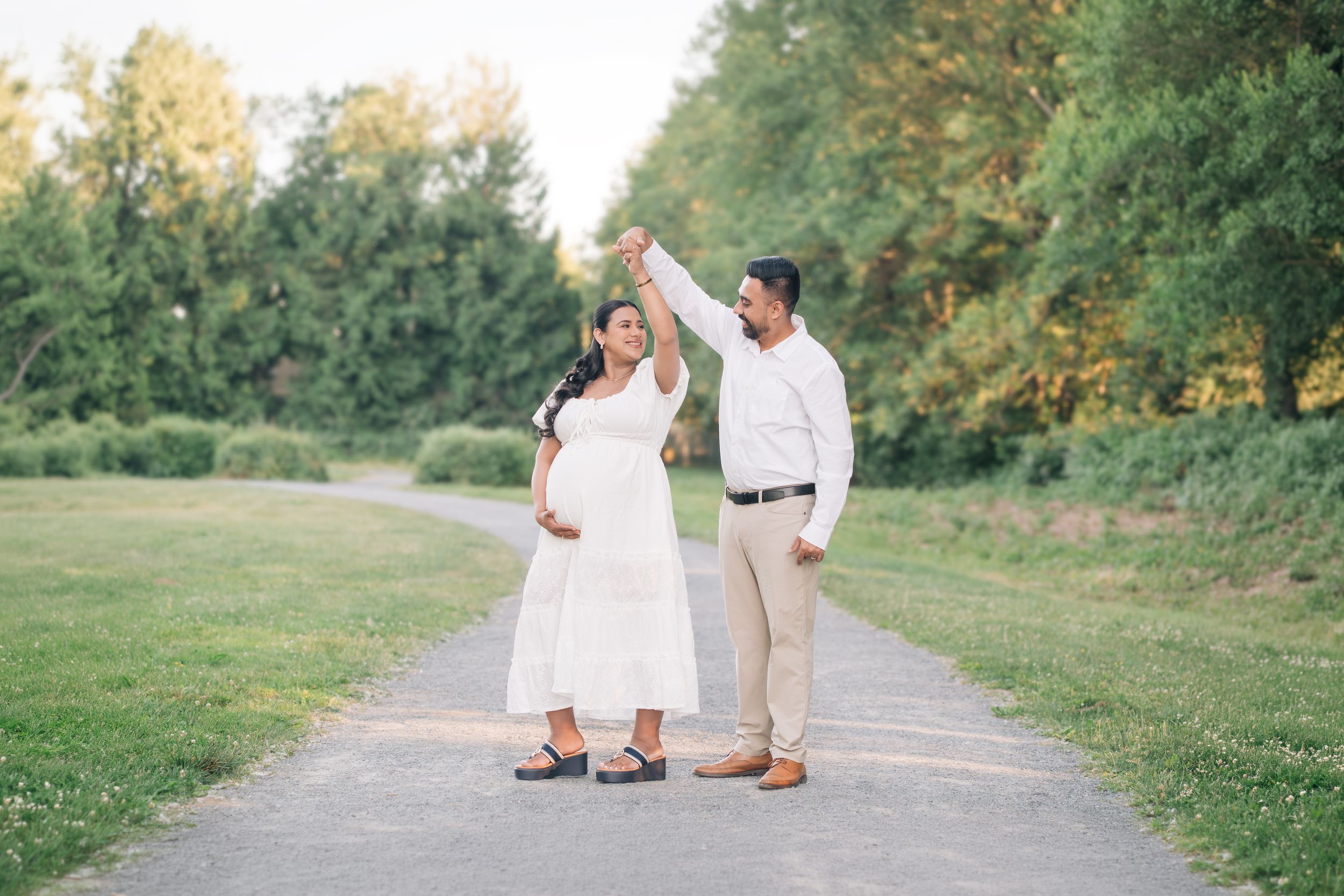 Pregnant woman in white dress dancing with man in white shirt on park path surrounded by lush green trees.