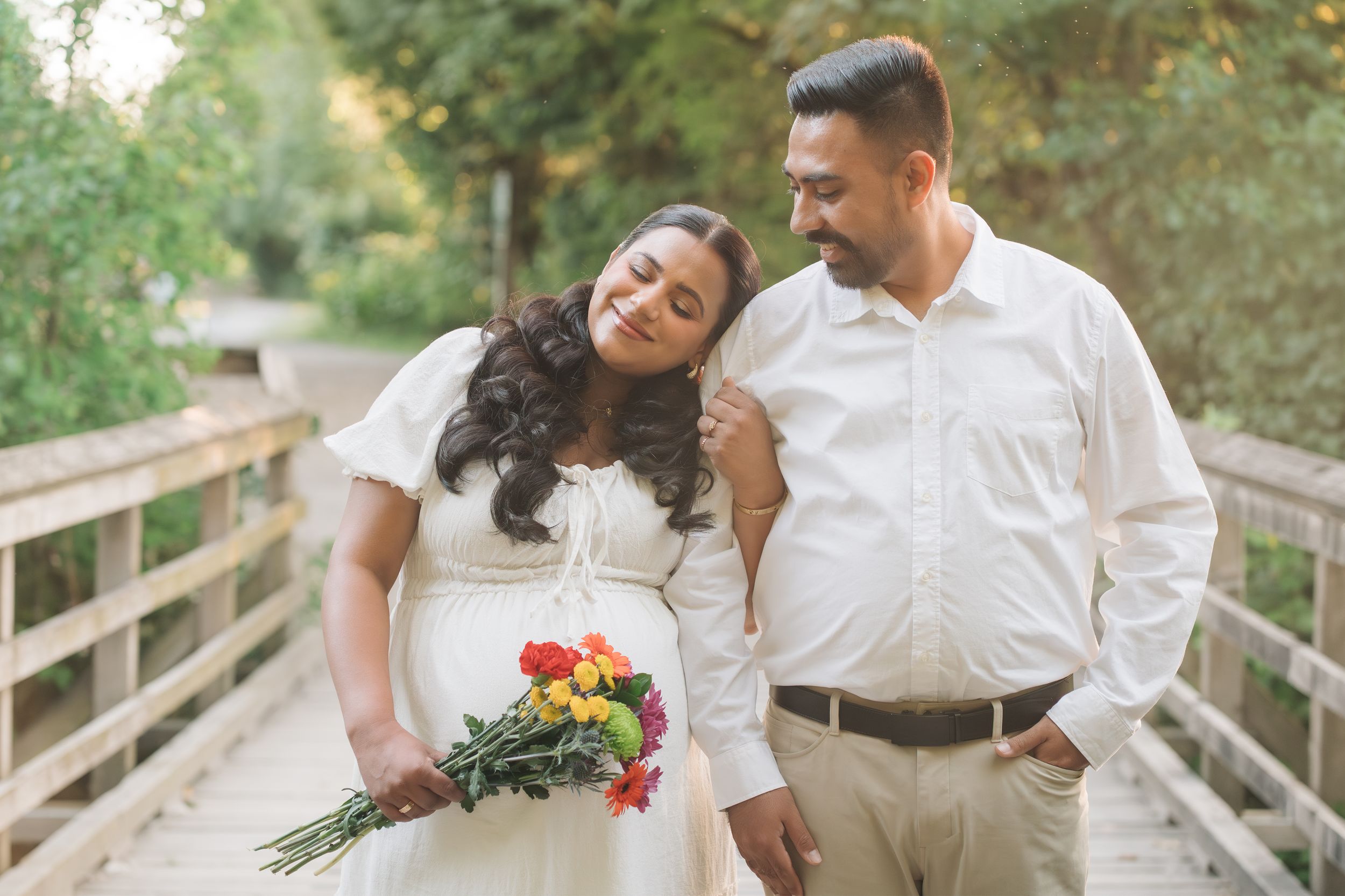 Pregnant couple in white attire on a wooden bridge, woman holding colorful bouquet, surrounded by lush greenery.