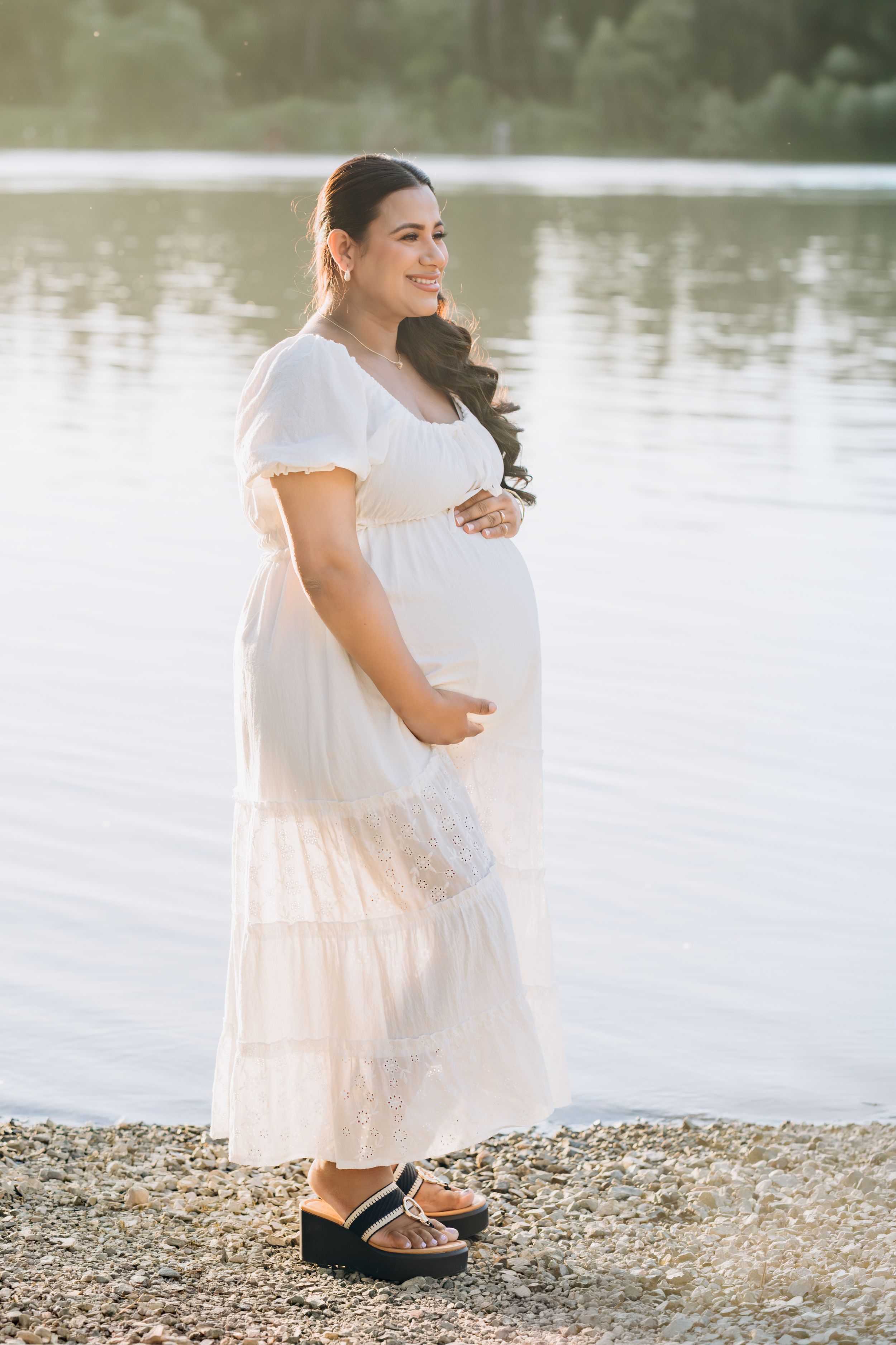 Pregnant woman in white maxi dress holding baby bump by a sunlit lake shore.
