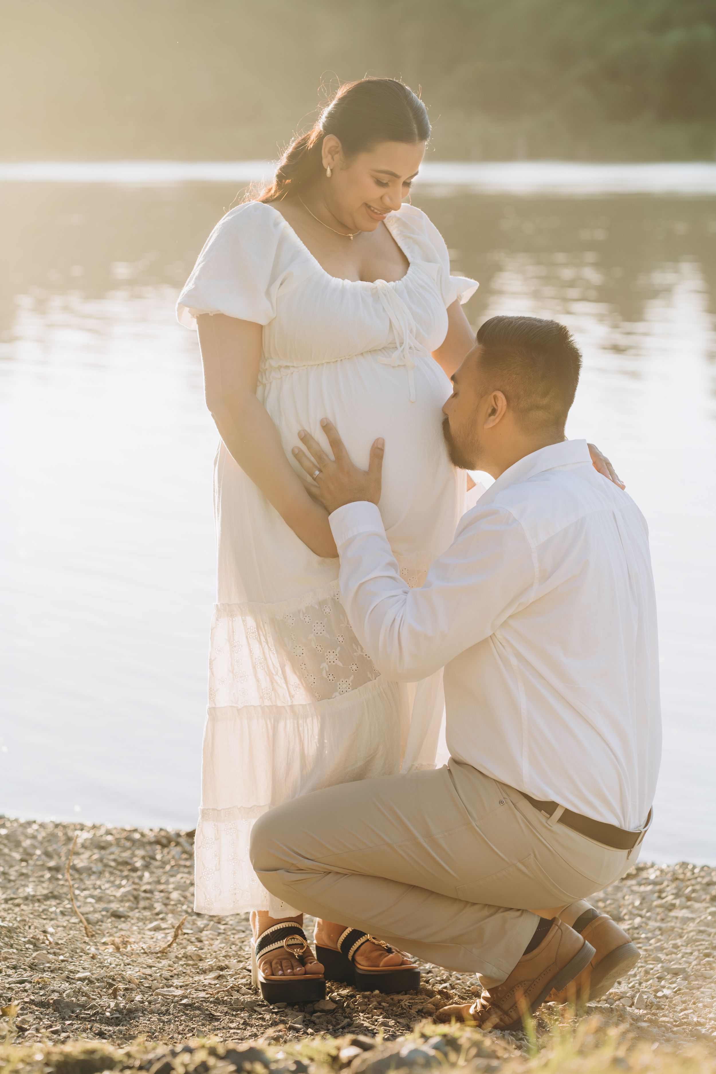 Man kissing pregnant womans belly outdoors by a lake at golden hour.