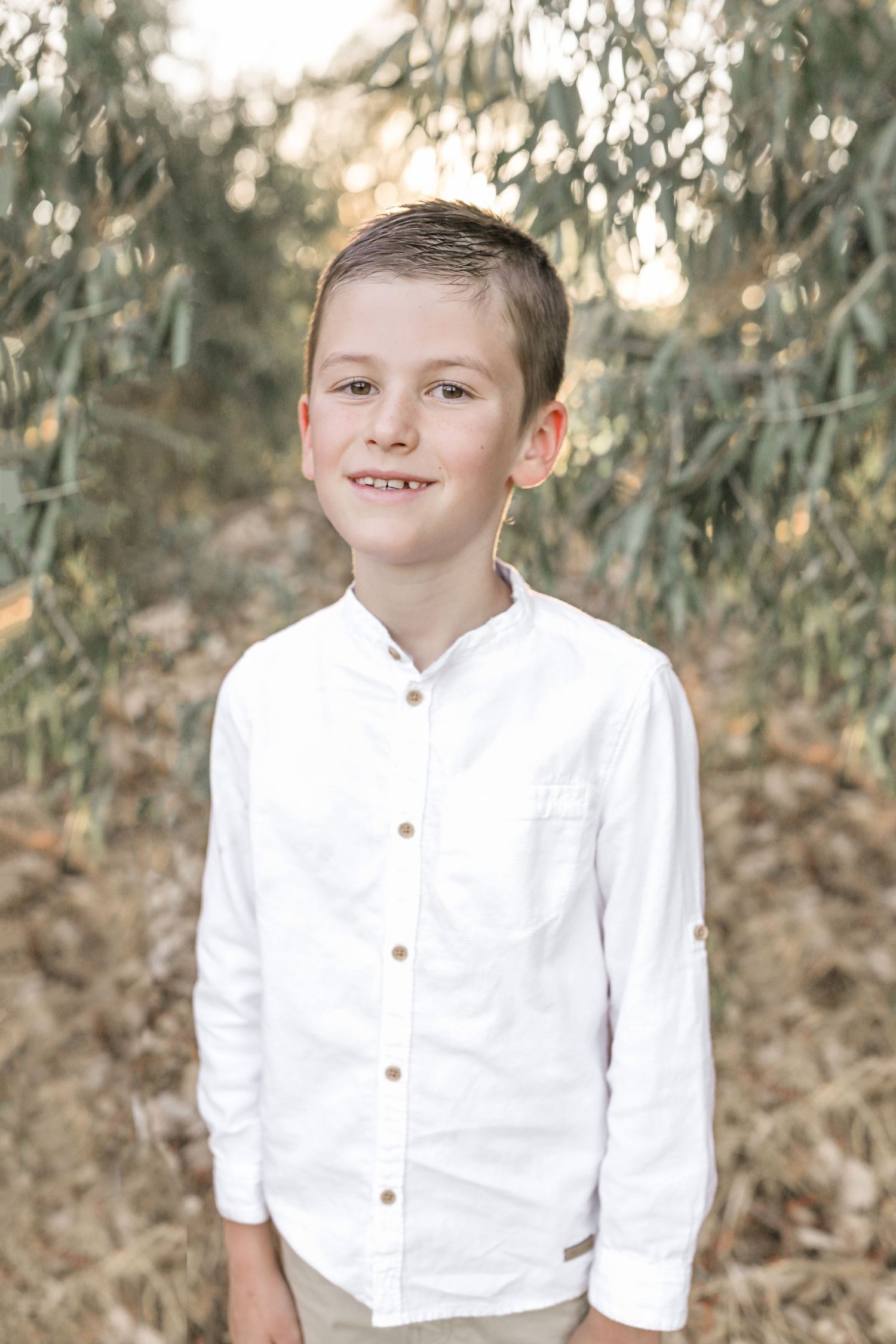 A smiling boy in a white button-up shirt stands outdoors among green foliage in a portrait photo.