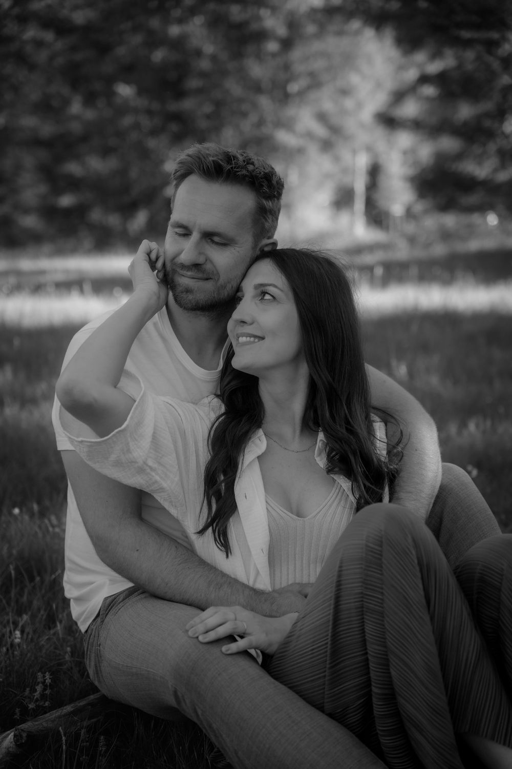 Black and white photo of a couple sitting outdoors, man embracing woman from behind as she smiles up at him.