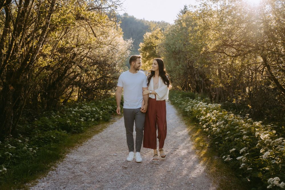 Couple holding hands walking on a sunlit gravel path surrounded by lush green trees and blooming flowers.