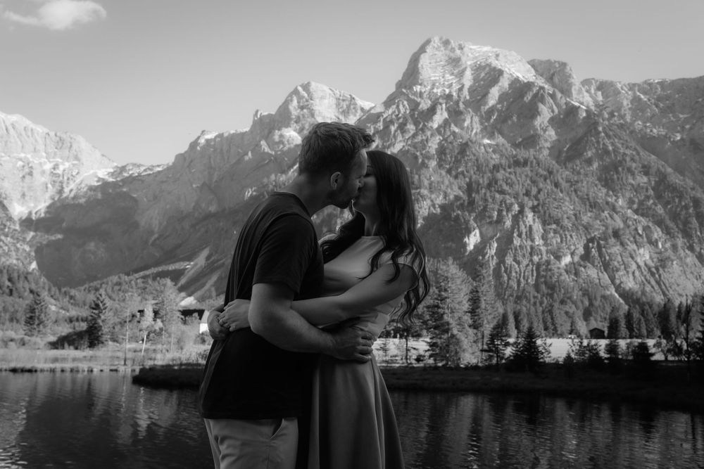 Couple sharing a romantic kiss with snow-capped Grand Teton mountains and lake in black and white photo.