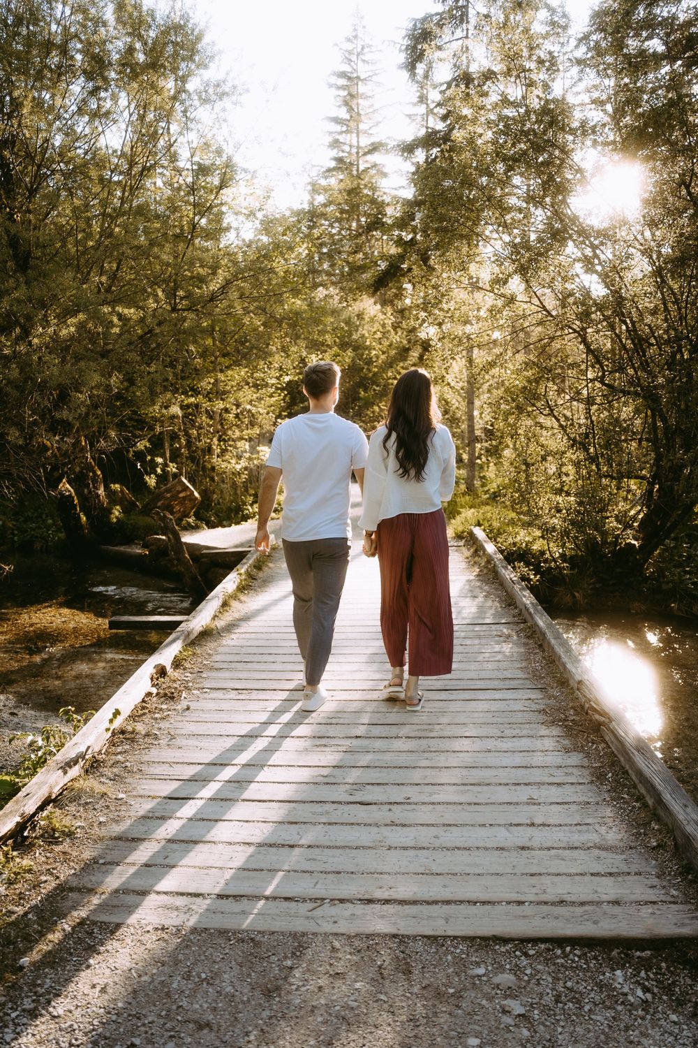 Couple holding hands walking on a wooden bridge surrounded by sunlit forest trees.