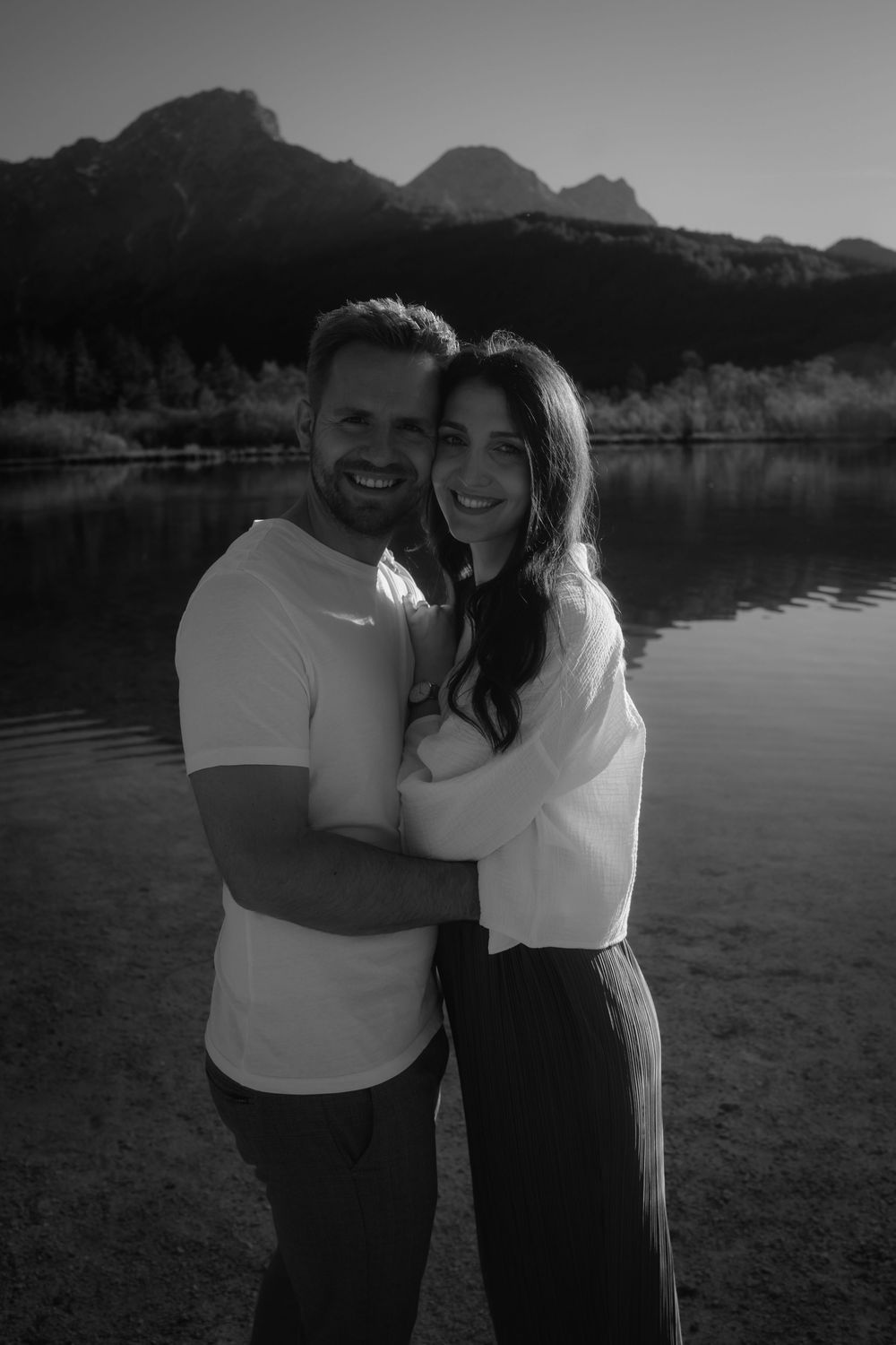 Black and white photo of a smiling couple embracing by a mountain lake with peaks reflected in the water.