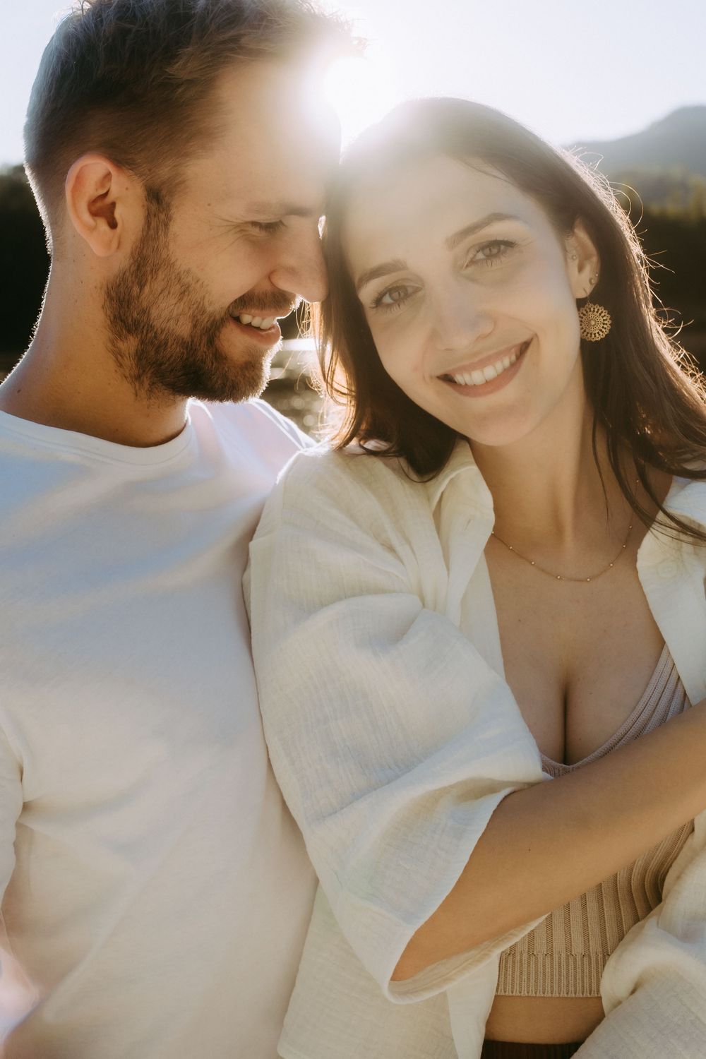 Couple embracing outdoors with sunlight glowing behind them, smiling warmly in white outfits near mountains.