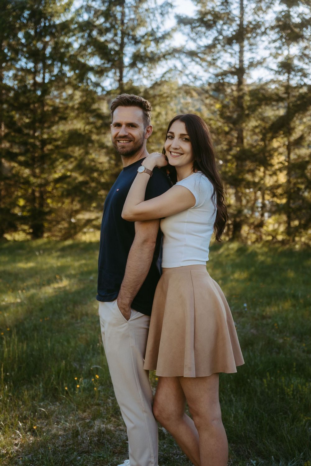 Couple smiling outdoors in a sunny forest, woman in white top and tan skirt embracing man in black shirt.