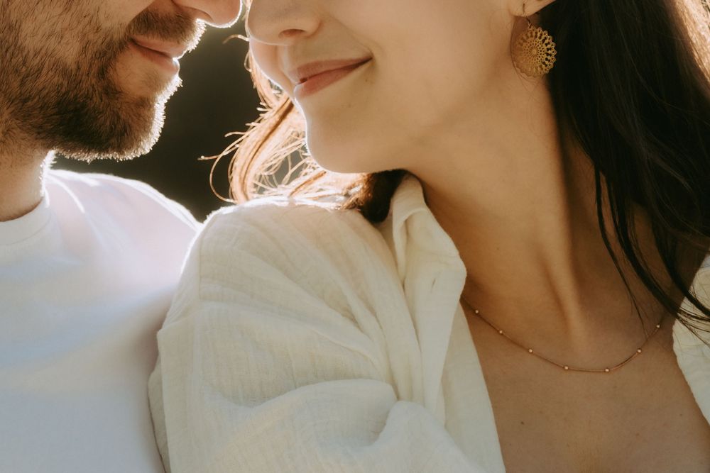 Romantic couple in white outfits sharing an intimate moment, woman wearing gold earring and necklace in warm sunlight.