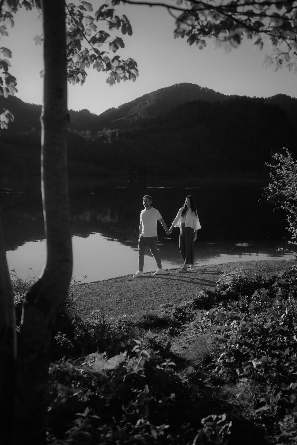 Black and white romantic couple holding hands by a reflective mountain lake surrounded by trees at dusk.