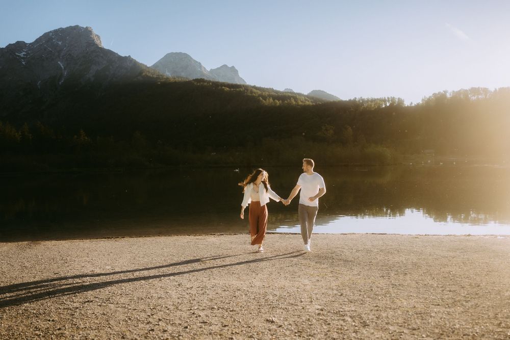 Couple holding hands walking along a serene mountain lake shore during golden hour sunset.