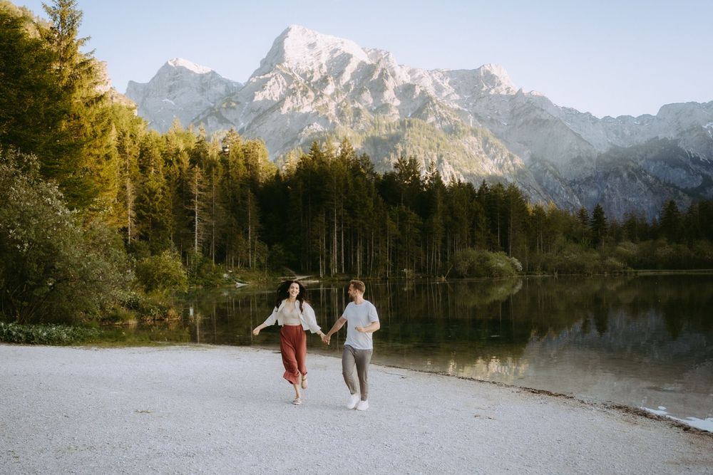 A joyful couple runs hand in hand along a lakeside path with stunning snow-capped mountains behind them.