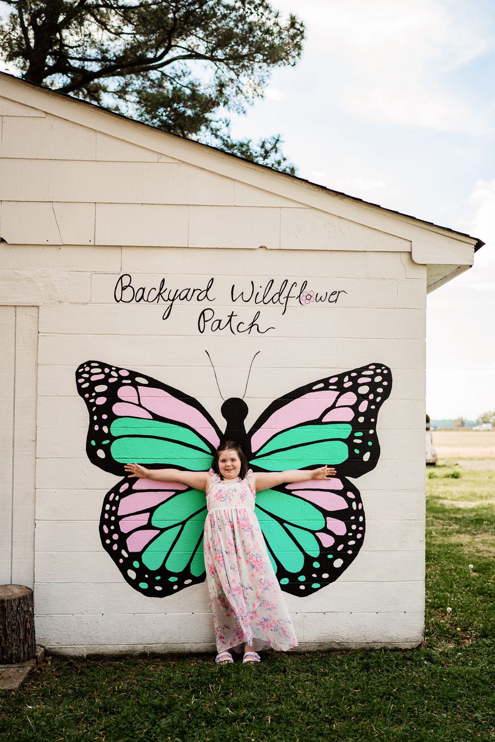 Young girl posing with colorful butterfly mural at Backyard Wildflower Patch, wearing pink floral dress.