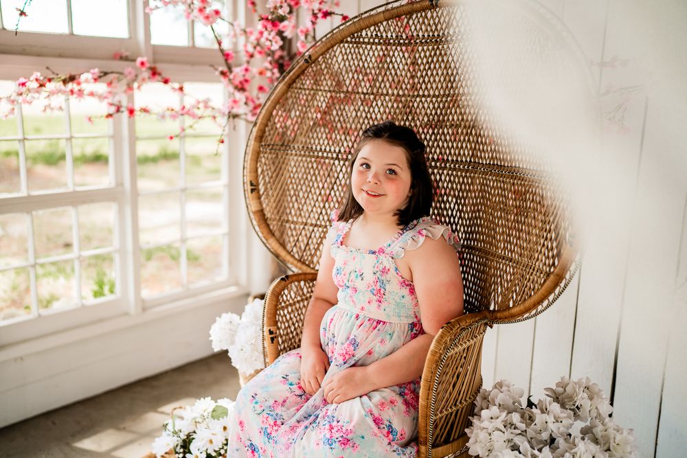 Young girl in floral dress seated in wicker peacock chair with cherry blossoms and white flowers in bright studio.