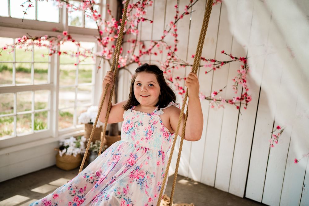 A young girl in a floral dress smiles on a rope swing with cherry blossom branches in a bright white room.