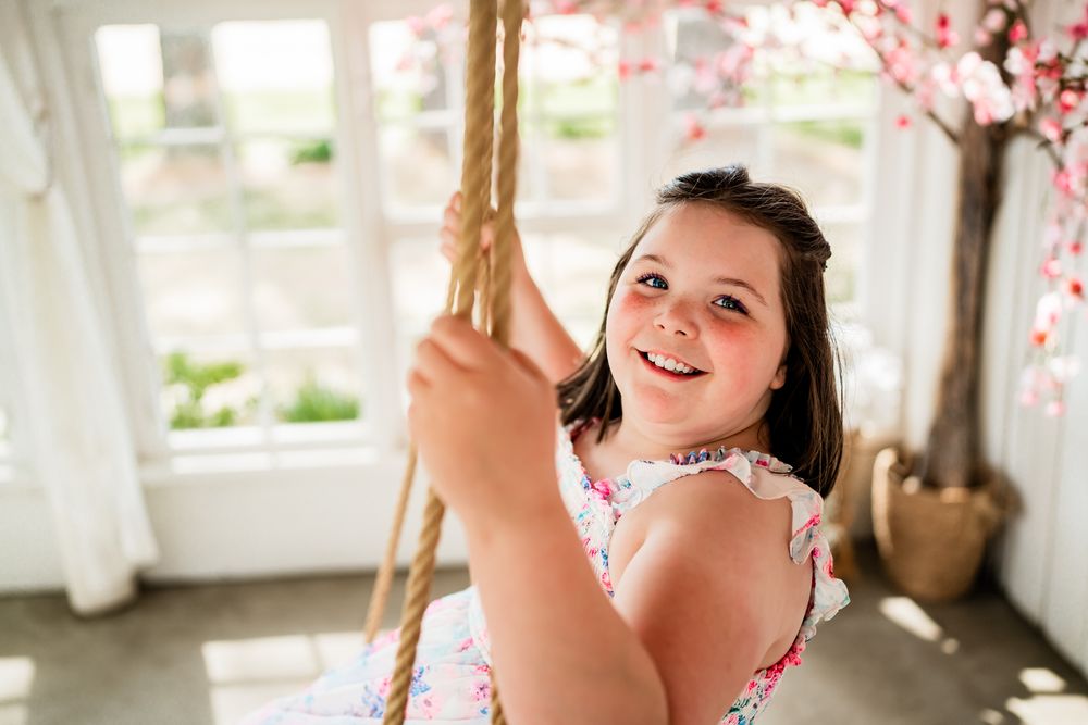 Smiling young girl on a rope swing indoors, wearing a floral dress with cherry blossom decor in background.