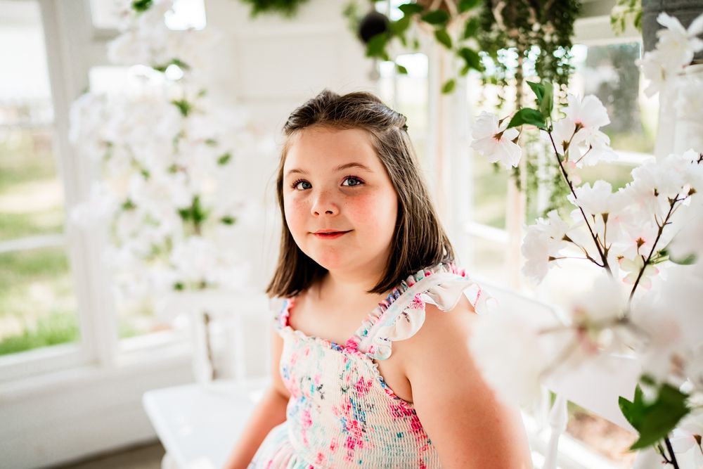 Young girl in floral dress smiling among white orchid blooms in a bright, flower-filled photography studio.