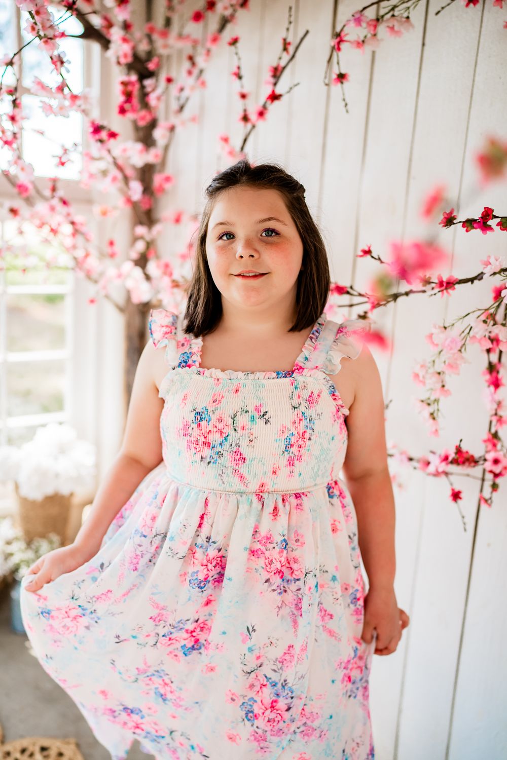 Young girl in floral dress posing among pink cherry blossom branches against white wooden backdrop.