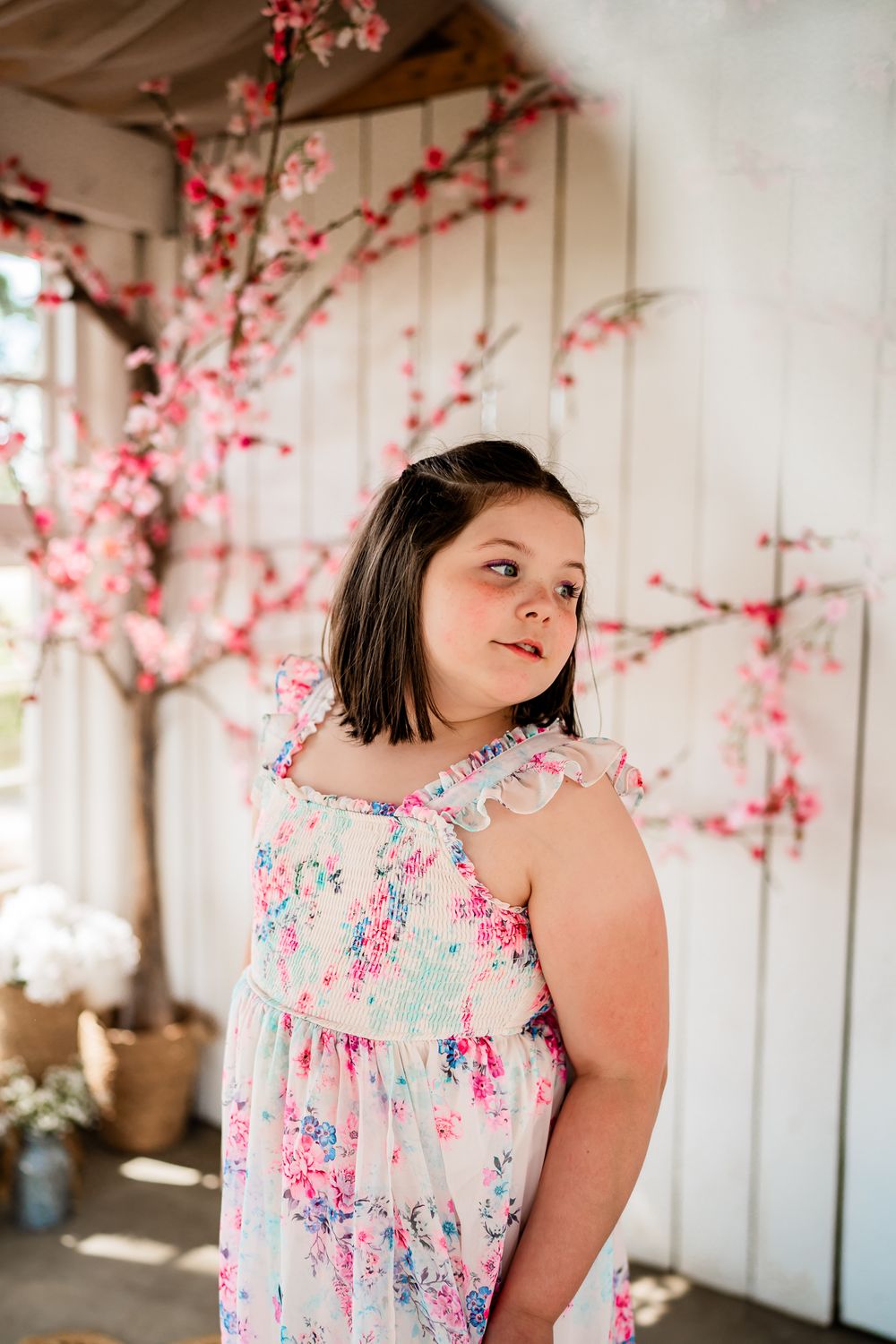 Young girl in floral sundress poses before white wall with pink cherry blossom branches in soft natural light.