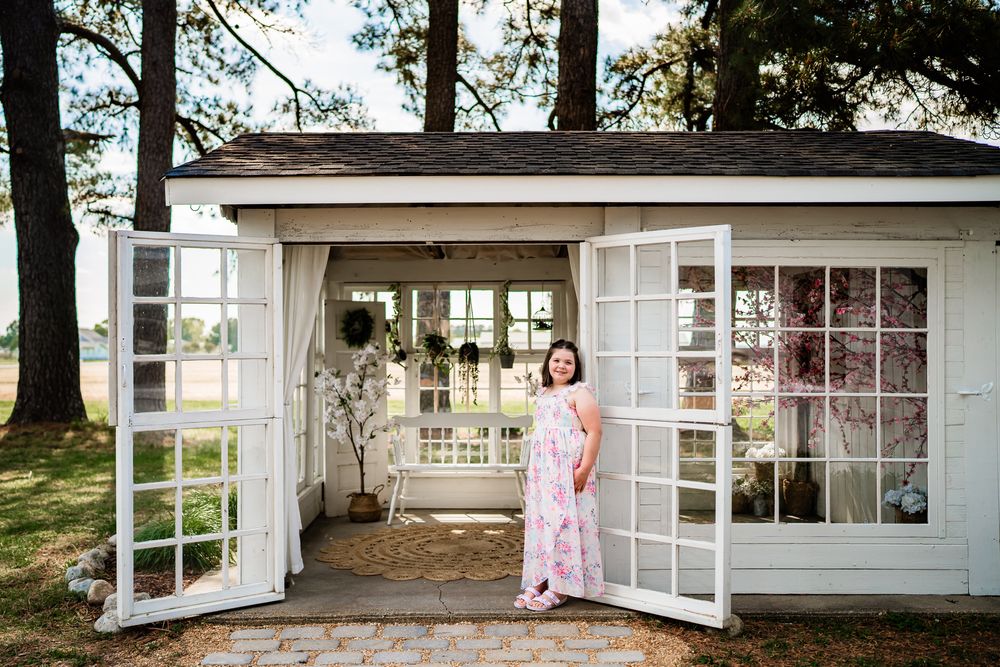 Woman in pink floral dress standing in doorway of charming white cottage with French doors outdoors.