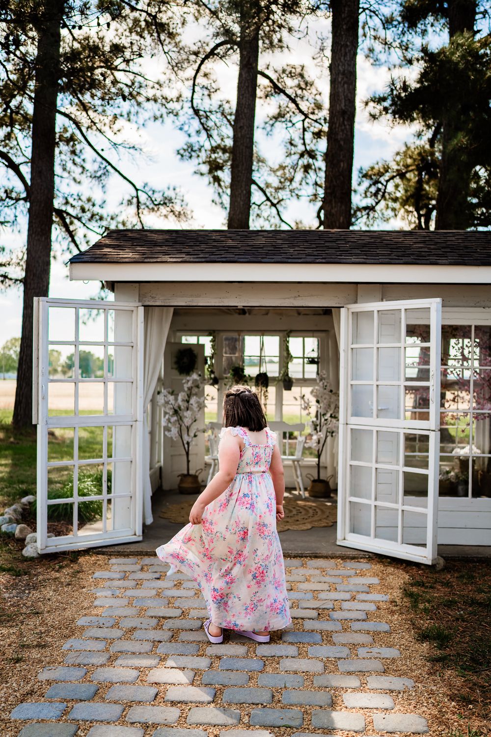 Woman in floral maxi dress walking into charming white shed with French doors on stone path.