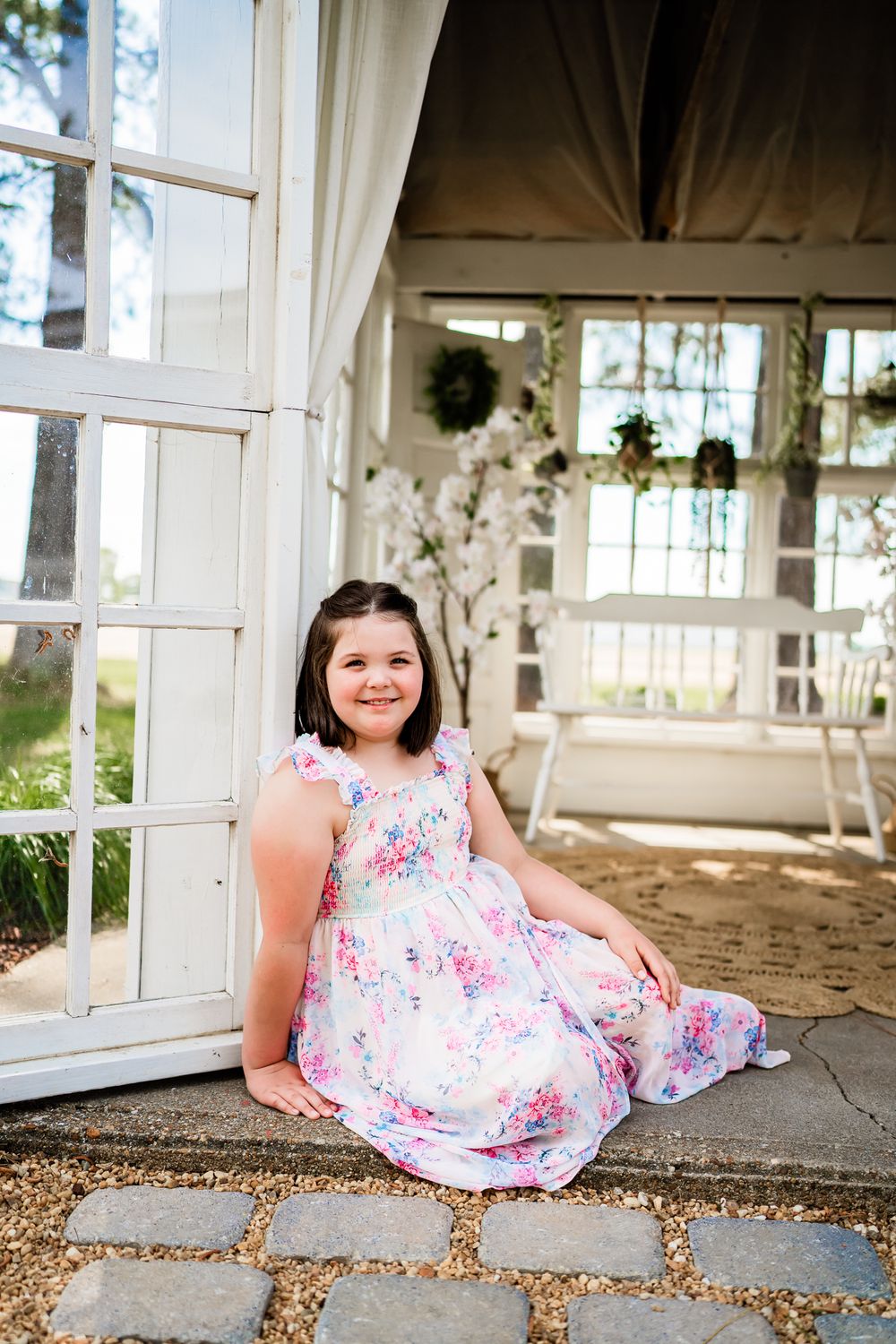 Young girl in floral dress sitting on stone steps beside white porch with bench and hanging plants.