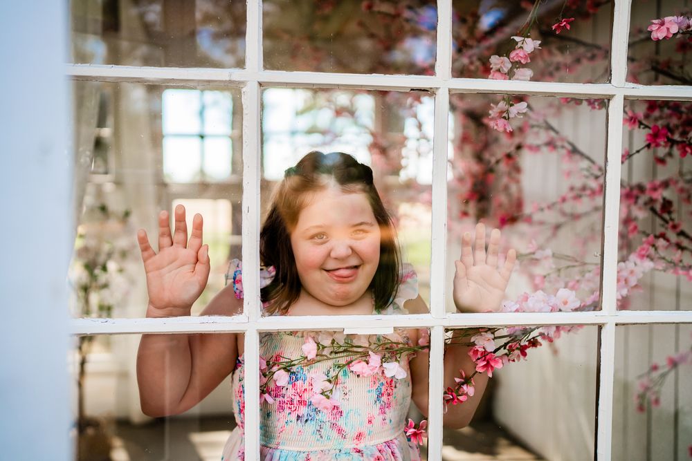 Young girl with Down syndrome waves through a white grid window surrounded by pink cherry blossoms in spring.