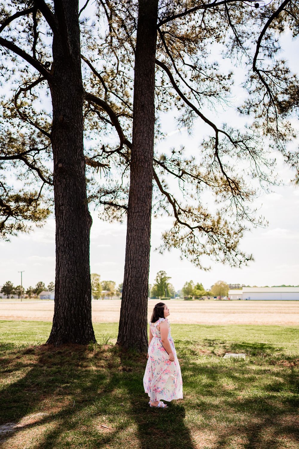 Young girl in floral dress standing between tall pine trees in a sunny park near a lake.