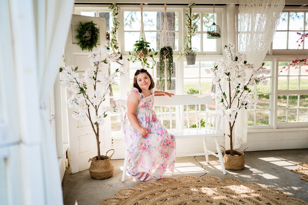 Woman in floral maxi dress posing on white swing bench in bright boho studio with cherry blossom trees.