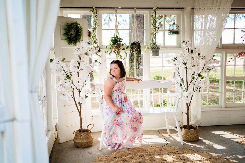 Woman in floral maxi dress posing by white bench in bright botanical studio with cherry blossom trees.