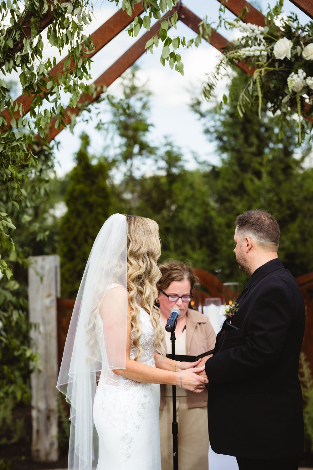 Bride and groom exchange vows outdoors under a wooden arch adorned with greenery and white flowers during a wedding ceremony.