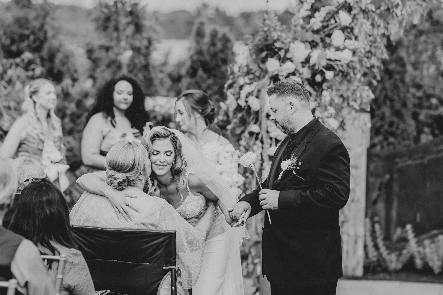 Black and white wedding ceremony photo of bride and groom exchanging rings outdoors with floral arch backdrop.