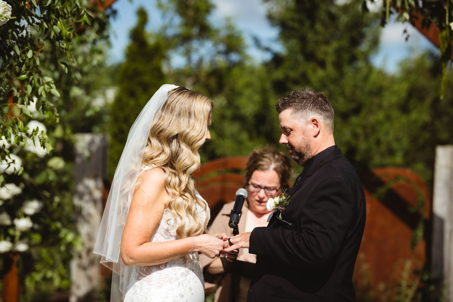 Bride and groom exchange rings during outdoor wedding ceremony with officiant and floral arch backdrop.