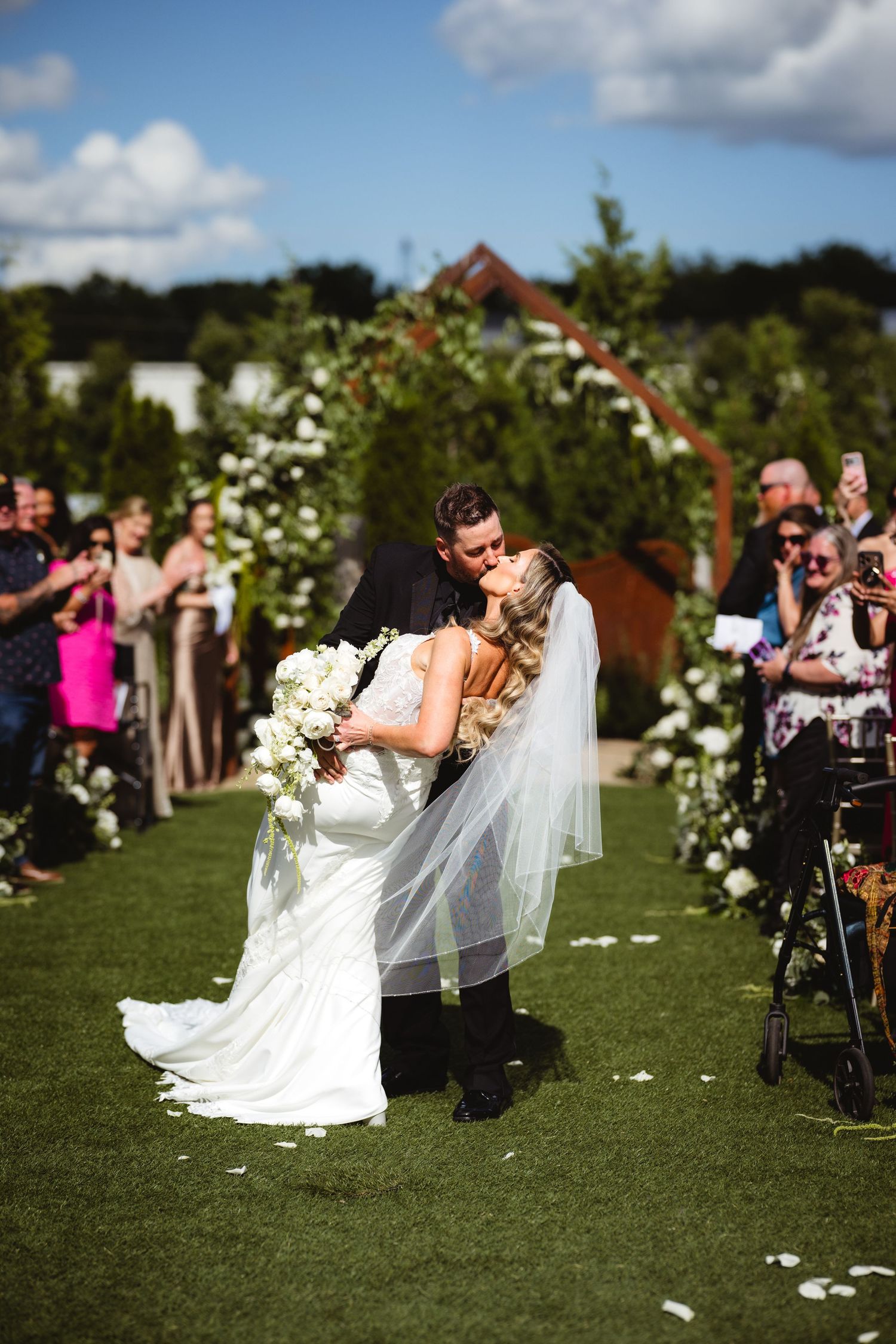 Bride and groom share first kiss at outdoor wedding ceremony on green lawn with floral arch and guests watching.