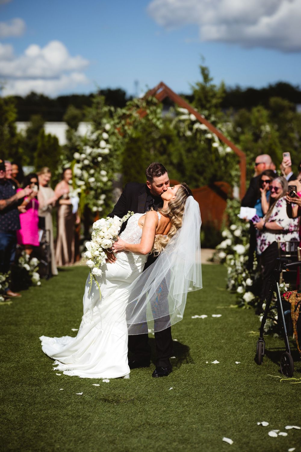 Bride and groom share first kiss at outdoor wedding ceremony on green lawn with floral arch and guests watching.