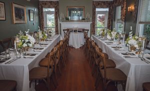 Elegant dining room set for a wedding reception with long white-draped tables, wooden chairs, floral centerpieces, and a fireplace.