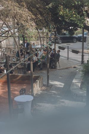 Guests seated outdoors under trees at a wedding ceremony venue with iron fence and patio seating.