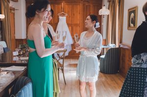 Two bridesmaids in green and white dresses toasting with champagne flutes near a hanging wedding gown indoors.