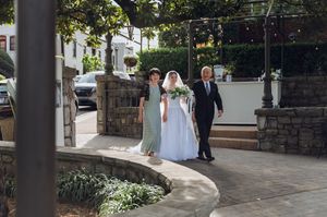 Bride walks with parents along stone pathway to wedding venue in elegant white gown.