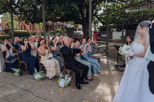 Wedding guests seated outdoors smile and photograph a bride in a white gown and veil holding a bouquet.
