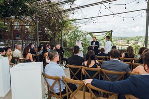 Outdoor wedding ceremony on a rooftop patio with string lights, wooden chairs, and guests watching couple exchange vows.