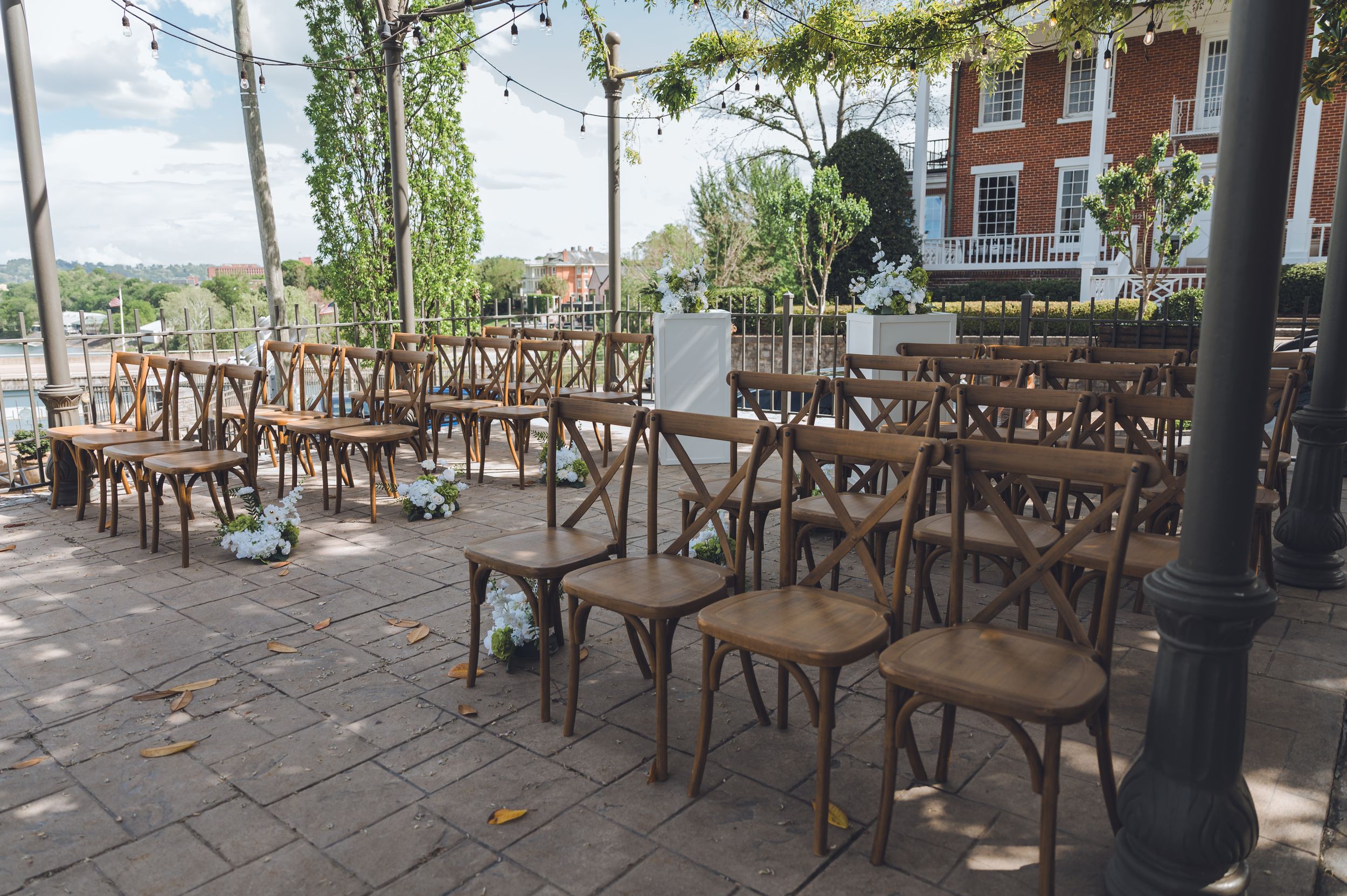 Rustic wooden cross-back chairs arranged for an outdoor wedding ceremony on a brick patio with lush greenery overhead.