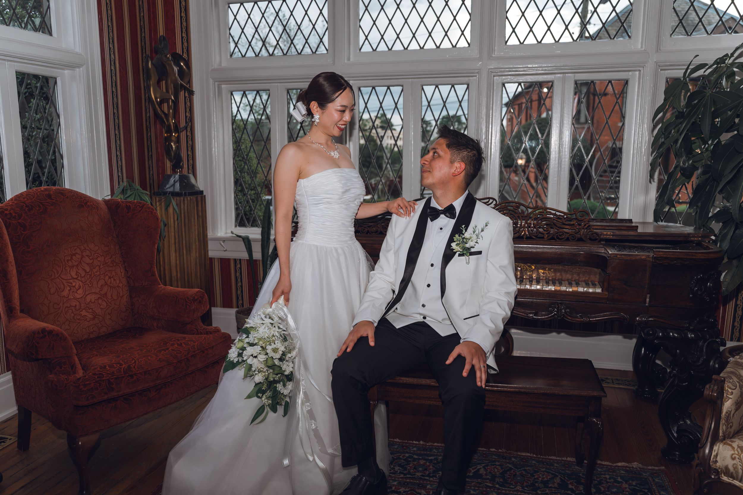 Bride in white strapless gown and groom in white tuxedo share a tender moment by a grand piano in an elegant room.